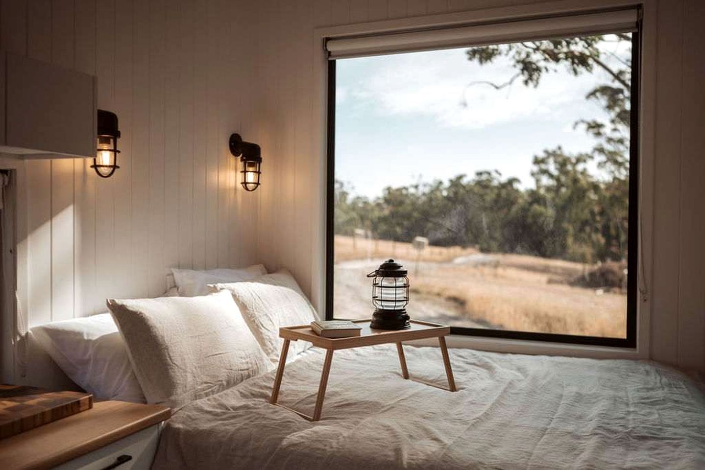 Un dormitorio con una cama, almohadas, una mesita de noche, un farol y vistas a un paisaje natural a través de un ventanal grande.