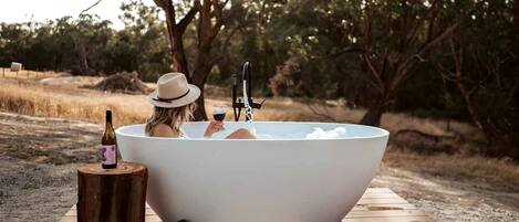 A person relaxing in a large outdoor bathtub with a bottle of wine and a glass of wine on a wooden platform.