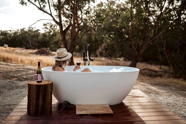 A person relaxing in a large outdoor bathtub with a bottle of wine and a glass of wine on a wooden platform.