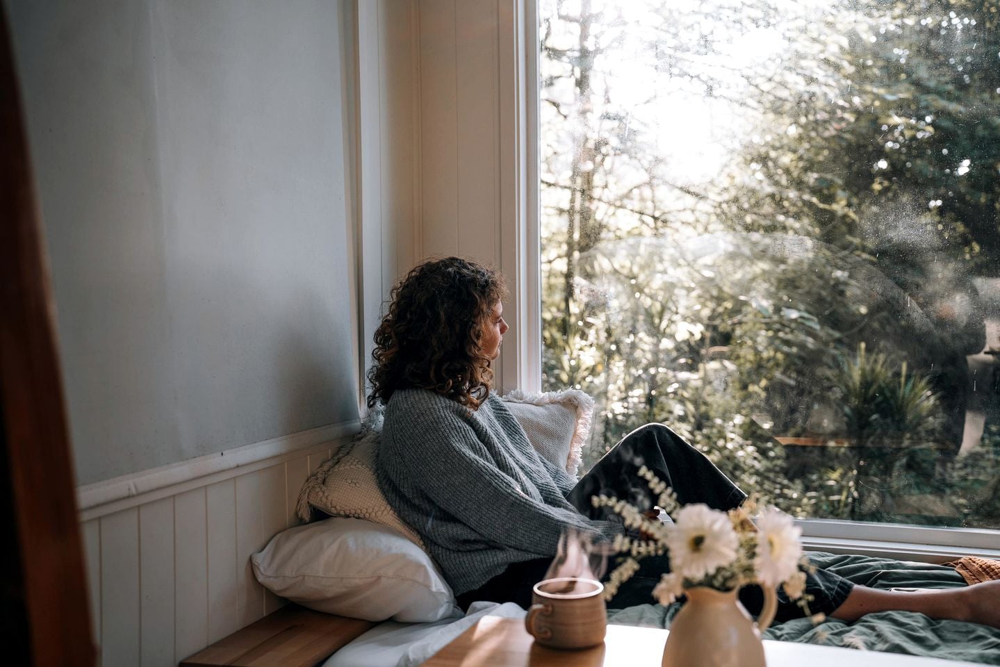 A person sitting by a window with a view of trees, a cushion, a mug, and a vase with flowers.