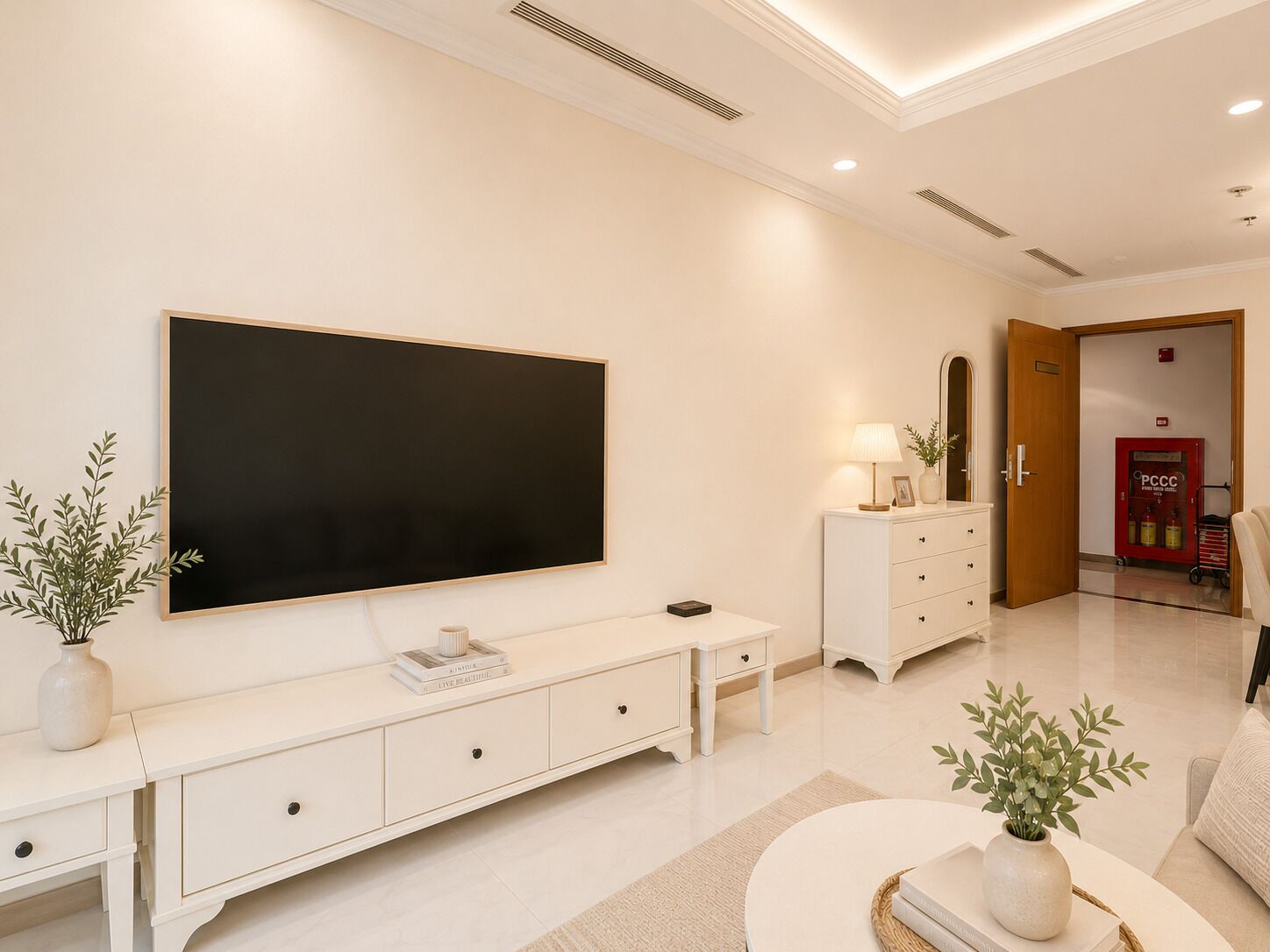 A modern living room with a large flat-screen TV, a white console table, a side table with a lamp, and a door leading to a hallway.