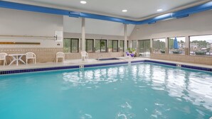 En indendørs swimmingpool med krystalklart blåt vand, hvide stole og udsigt til bygninger gennem store vinduer.