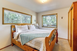 A bedroom with a wooden bed, two windows with views of trees, a bedside table with a lamp, and a door to the right. - Deck & Mtn Views! Woodsy Haven in Running Springs (Running Springs)