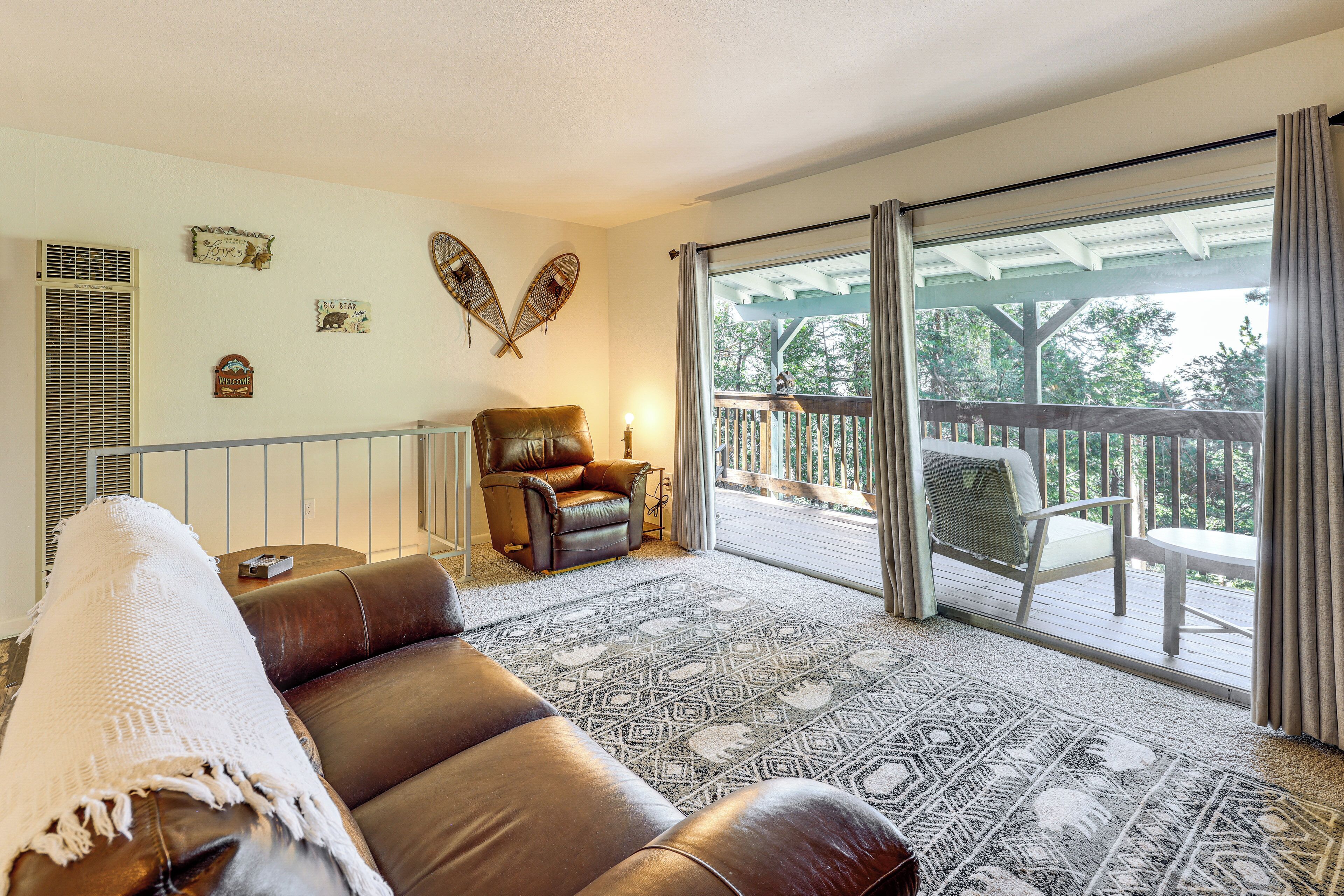 Ein Wohnzimmer mit braunem Ledersofa, einem gemusterten Teppich, einem Holzstuhl und einem Balkon mit Blick auf Bäume.