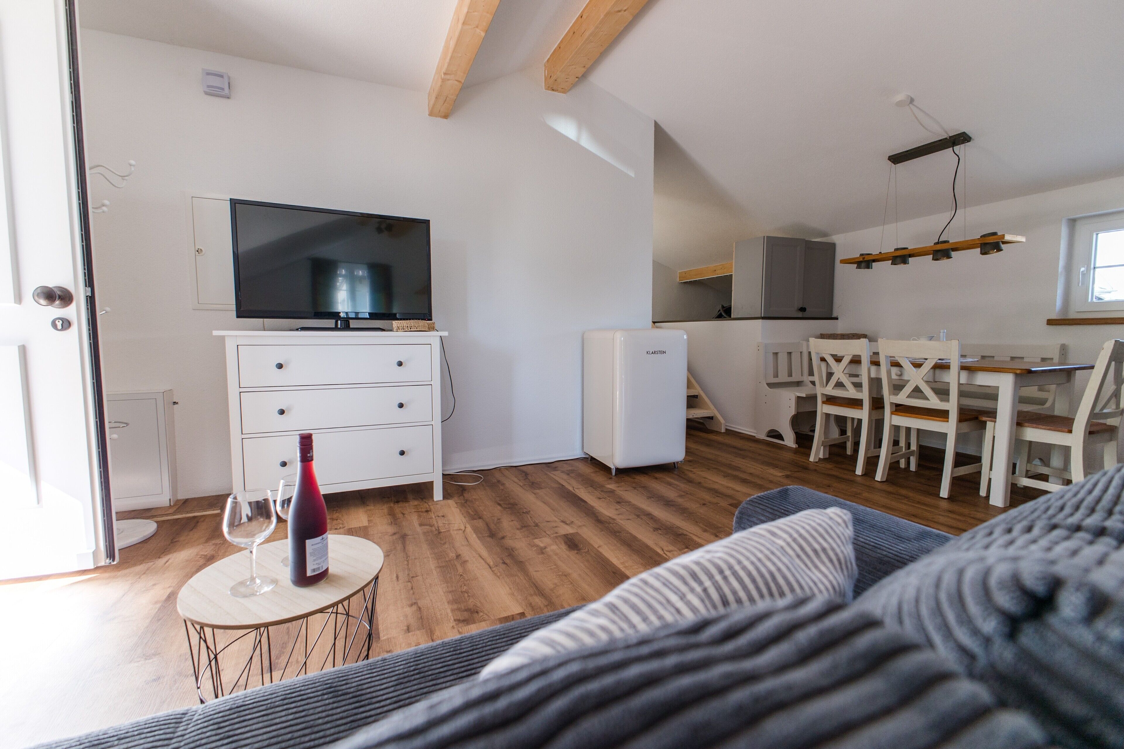Ein kompakter Wohnbereich mit Sofa, einem kleinen runden Tisch mit Weinflasche und Glas, einem Fernseher auf einer Anrichte, einer Essecke mit Stühlen und Tisch sowie einer Küchenzeile mit Schränken und Kühlschrank.