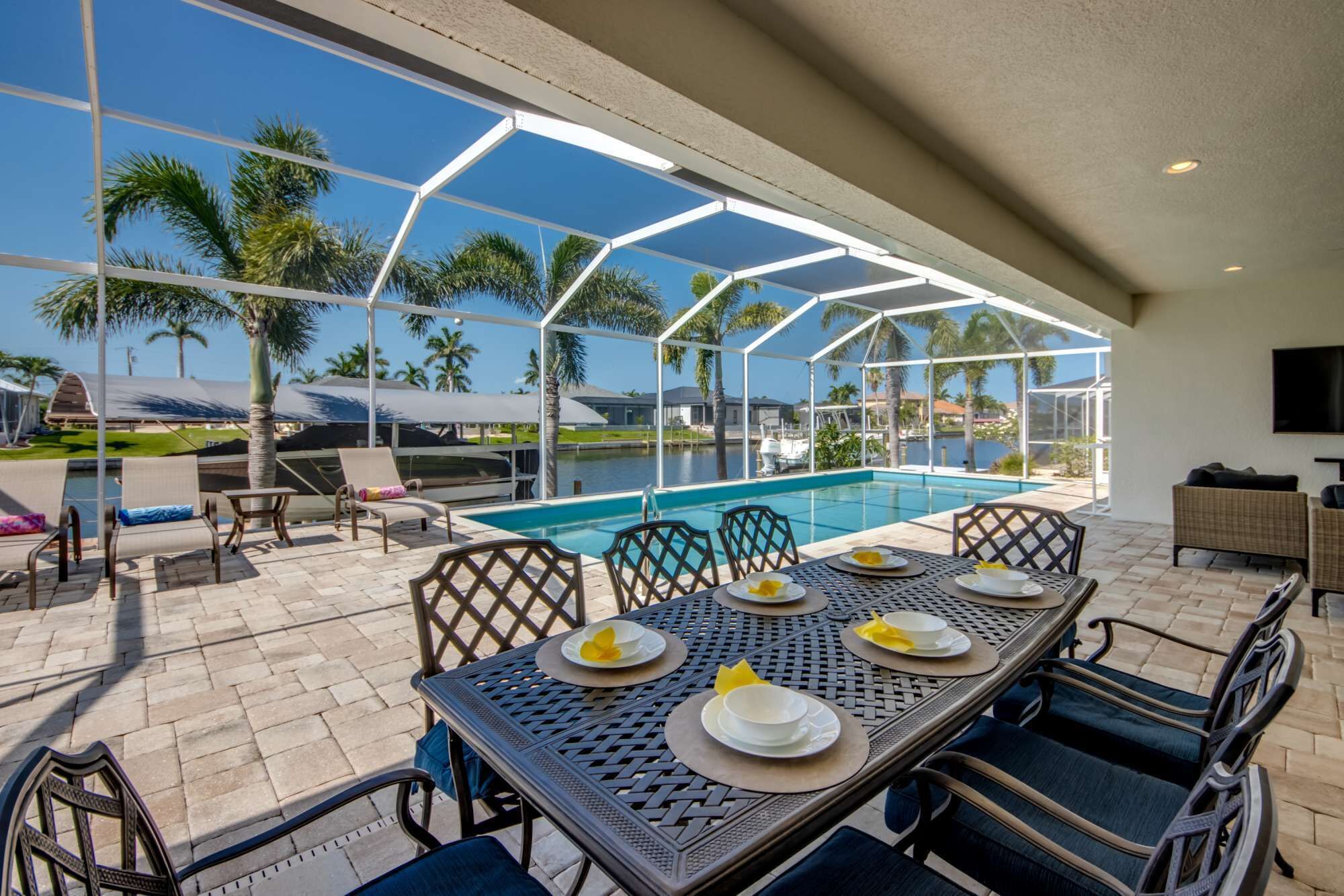 Un área de comedor junto a la piscina con una mesa preparada para cuatro, sillas y vistas a la piscina y palmeras.