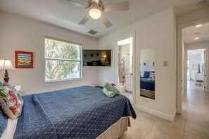 Ein Schlafzimmer mit einem Bett, einem Deckenventilator, einem Fenster, einem Spiegel und einem durchgehenden Türspalt sichtbaren Badezimmer.