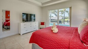 Un dormitorio con una cama grande, un televisor sobre un tocador blanco y vista a una piscina por medio de un ventanal.
