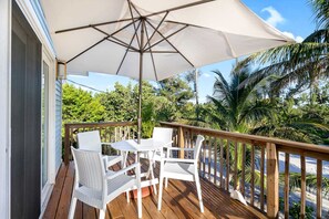 Eine Holzterrasse mit Tisch und Stühlen, beschattet von einem großen Sonnenschirm, umgeben von Palmen und einem klaren Himmel.