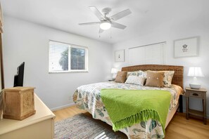 Ein Schlafzimmer mit einem Bett, einem Deckenventilator, zwei Nachttischen mit Lampen, einem Holznachttisch und einem Fenster mit Jalousien.