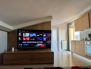 Ein modernes Wohnzimmer mit großem Flachbildfernseher, der eine Streaming-Oberfläche anzeigt, Holzschränken und Blick zur Küche.