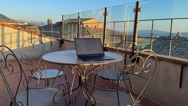 A balcony with a table and chairs, a laptop, and a view of buildings and hills. - Luce, Penthouse with Panoramic Terrace (Assisi)
