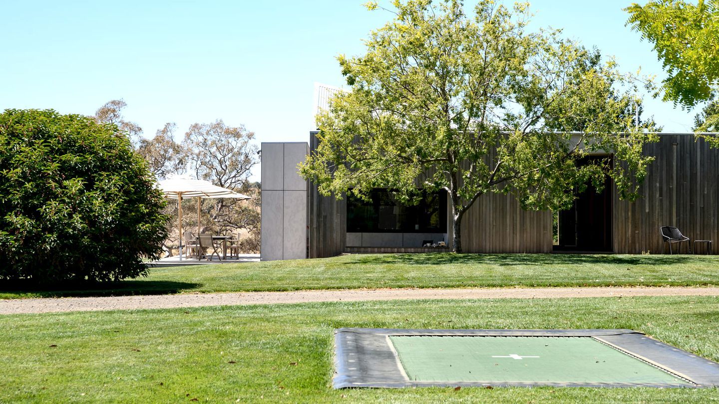 A grassy area with a trampoline, a wooden fence, and a building with a large window.