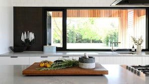 A modern kitchen with a large window, a wooden cutting board with fresh produce, a mortar and pestle, and a knife block.