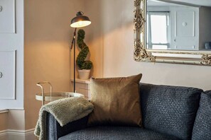 A dark-colored sofa with a textured fabric, a single decorative pillow, a small potted plant, a floor lamp, and a mirror with an ornate frame. - Host & Stay - Captain's Quarters (Seaton Carew)
