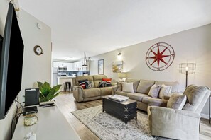 Ein Wohnzimmer mit einer Couch, einem Couchtisch, einem Fernseher, einer Küche im Hintergrund und einer großen Wanduhr.