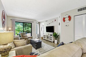 A living room with a sofa, armchair, TV, and a large window. - Ocean Oriented Oasis!  Indoor Heated Pool!  Pickleball! Hot Tub! (Hilton Head)