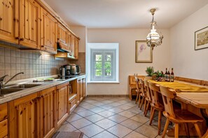 Een keuken met houten kastjes, een betegelde spatwand, een raam, een eettafel met stoelen en een kroonluchter.