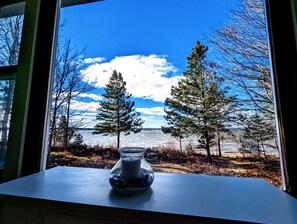 A table with a candle and a vase, overlooking a lake with trees and a clear sky. - Serene Waterfront Beach House with Private Shoreline & Fire Pit Near Acadia National Park | Trenton, Maine (Trenton)
