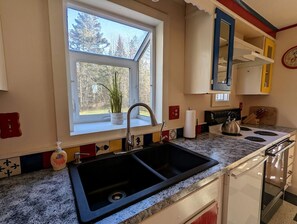 A kitchen with a double sink, a stove, and a window with a view of trees. - Serene Waterfront Beach House with Private Shoreline & Fire Pit Near Acadia National Park | Trenton, Maine (Trenton)