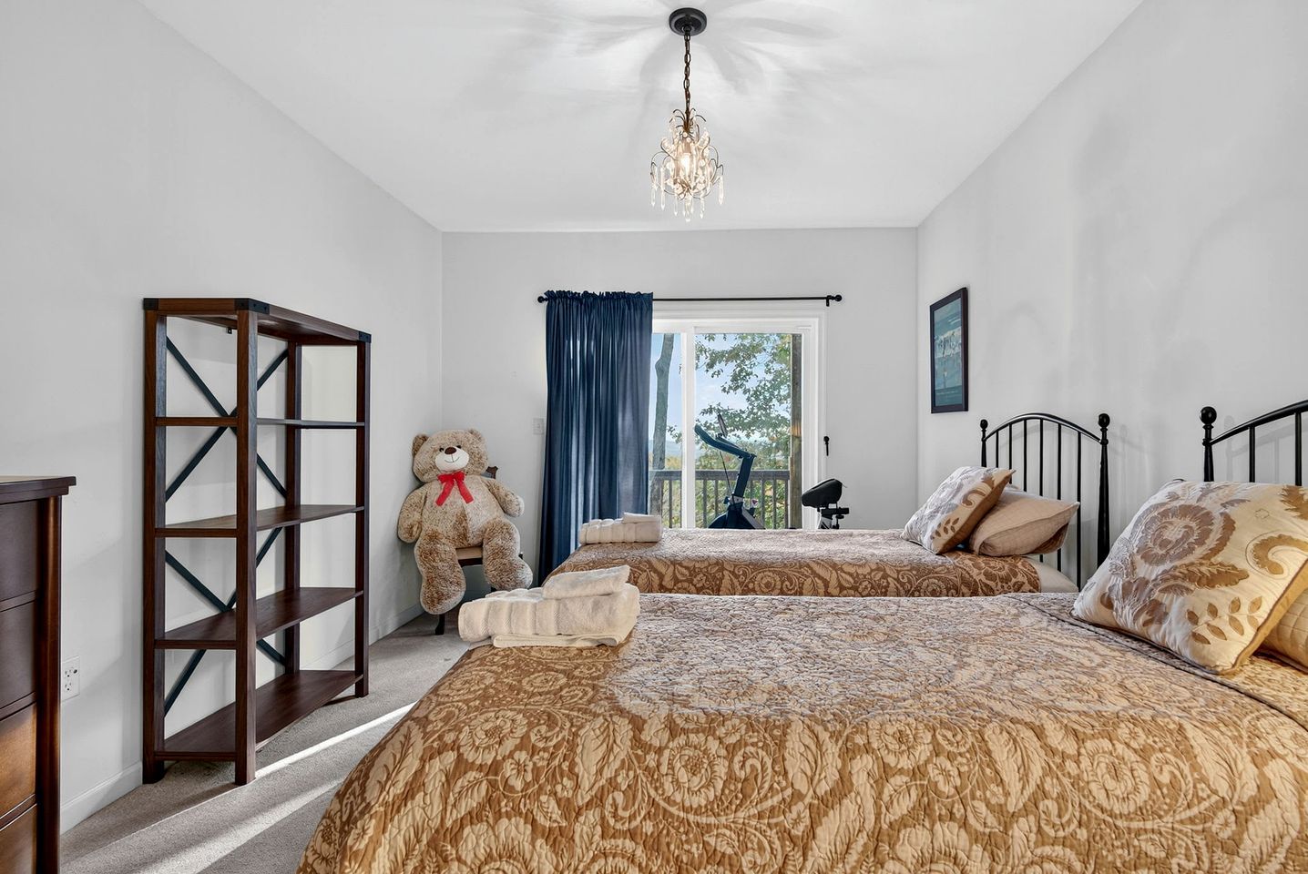 Une chambre avec deux lits, un grand ours en peluche, un lustre et une vue sur l’extérieur.