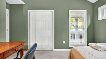 A bedroom with a bed, a desk, and a chair. There is a white door and a window with blinds.
