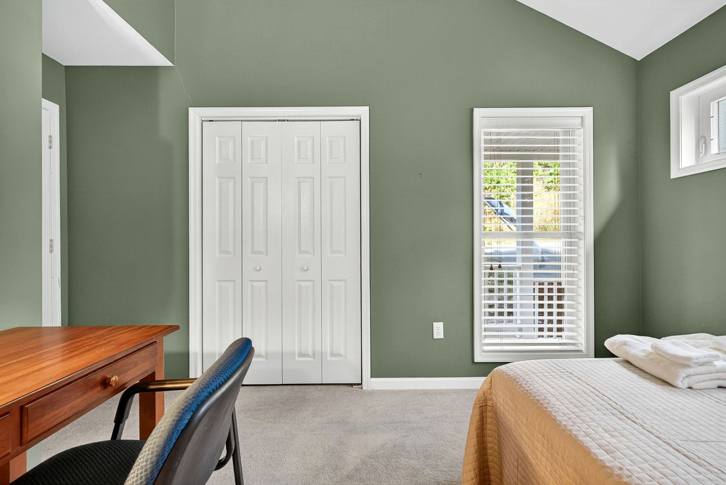 A bedroom with a bed, a desk, and a chair. There is a white door and a window with blinds.