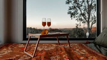 A bed with a floral bedspread, a wooden bedside table with two glasses of a beverage and a flower, and a view of trees through a window.