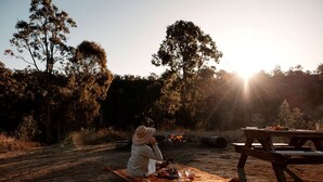 Una persona sentada sobre una manta al aire libre, con una mesa de picnic y un fuego al fondo.