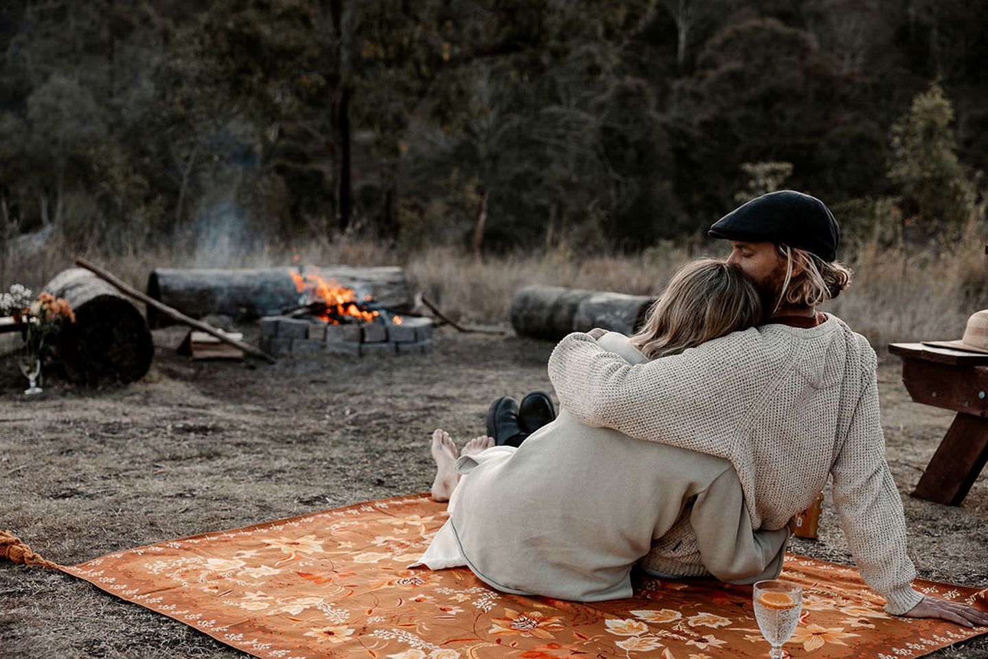 Two people sitting on a blanket outdoors with a campfire in the background.