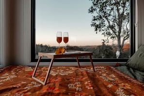 Un lit recouvert d’une couvre-lit à motifs floraux, une table de chevet en bois avec deux verres contenant une boisson et une fleur, et une vue sur les arbres par la fenêtre.