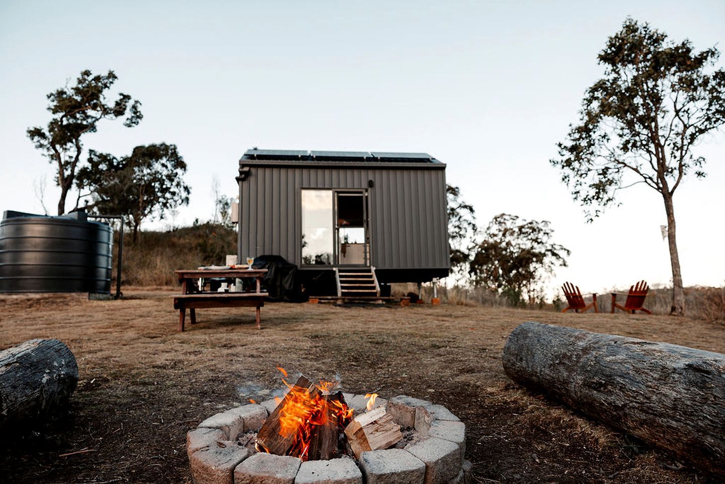 A modern cabin with a fire pit, outdoor seating, and a water tank in a rural setting.