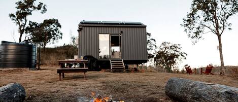 A modern cabin with a fire pit, outdoor seating, and a water tank in a rural setting.