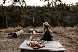 Une personne est assise à une table de pique-nique en bois, à l’extérieur, avec un foyer au fond.