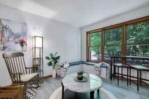 A modern interior with a dining area, a large window with a view of trees, a bookshelf, and a painting on the wall. - Sandlewood Getaway  Fire Pit  Pool  Hot Tub (East Stroudsburg)