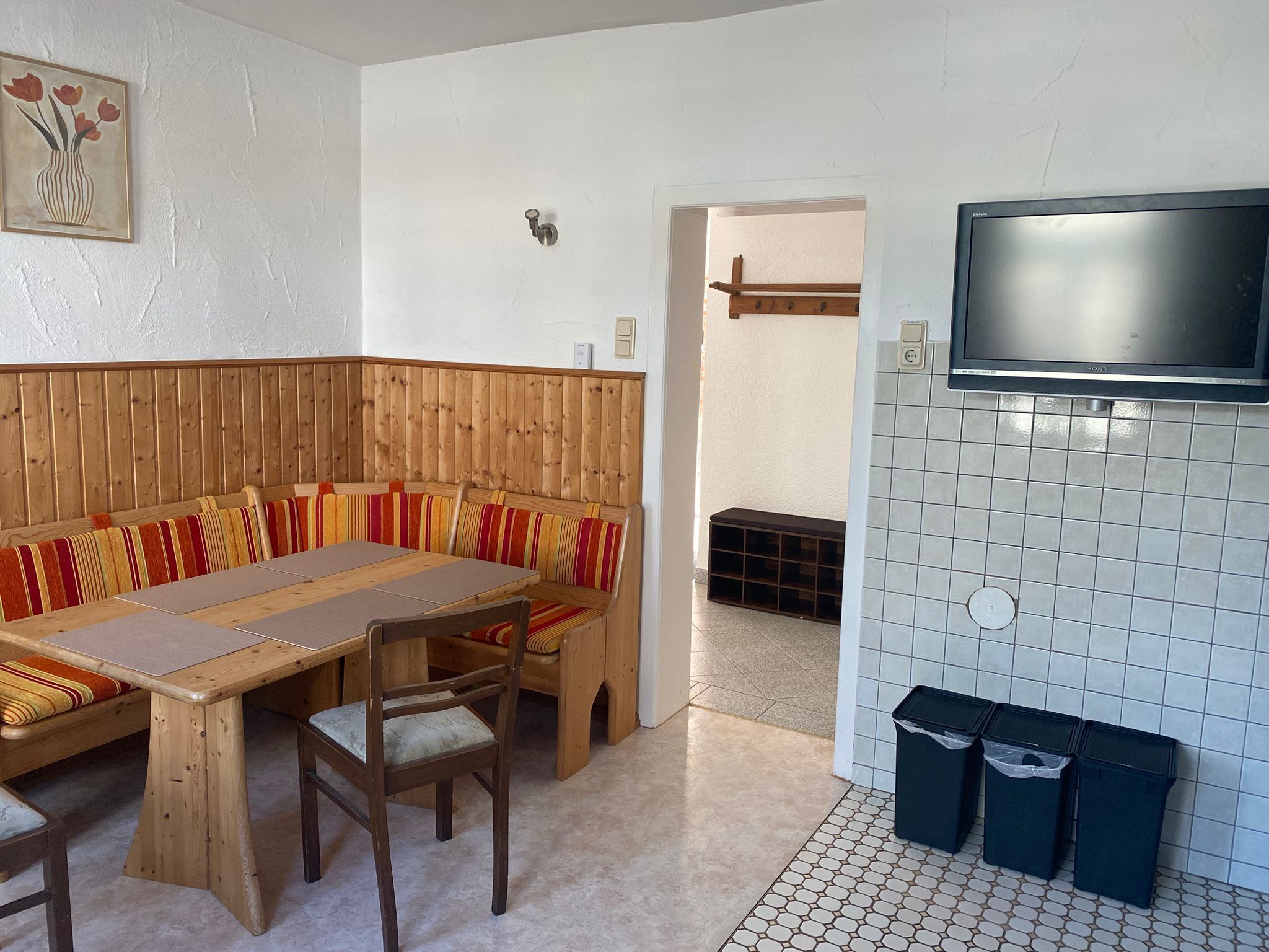 A room with a wooden bench, a table, a TV mounted on a tiled wall, and trash bins.