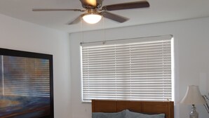 A bedroom with a bed, a ceiling fan, a window with blinds, a bedside table with a lamp, and a painting on the wall. - 2-Min Walk to Beach · 1BR Duplex w/ Private Lot (Redington Shores)