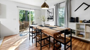 Uma área de refeições com mesa de madeira, cadeiras pretas, luminária pendente e uma estante com livros e objetos decorativos.