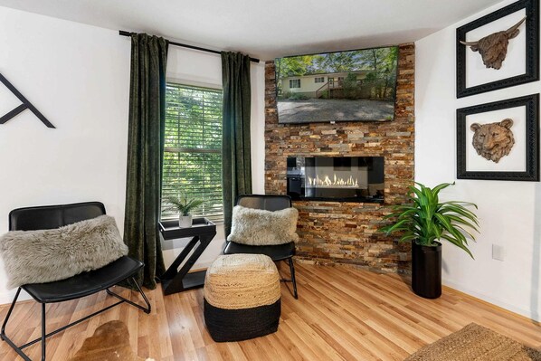A living room with a fireplace, two chairs, a sofa, a potted plant, and a window with curtains.