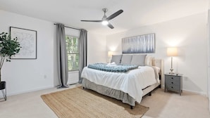 Um quarto com uma cama, um ventilador de teto, uma mesa de cabeceira, uma luminária, um quadro emoldurado e uma janela com cortinas.