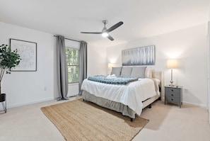 Una camera da letto con un letto, una ventola a soffitto, un comodino, una lampada, un quadro incorniciato e una finestra con tende.