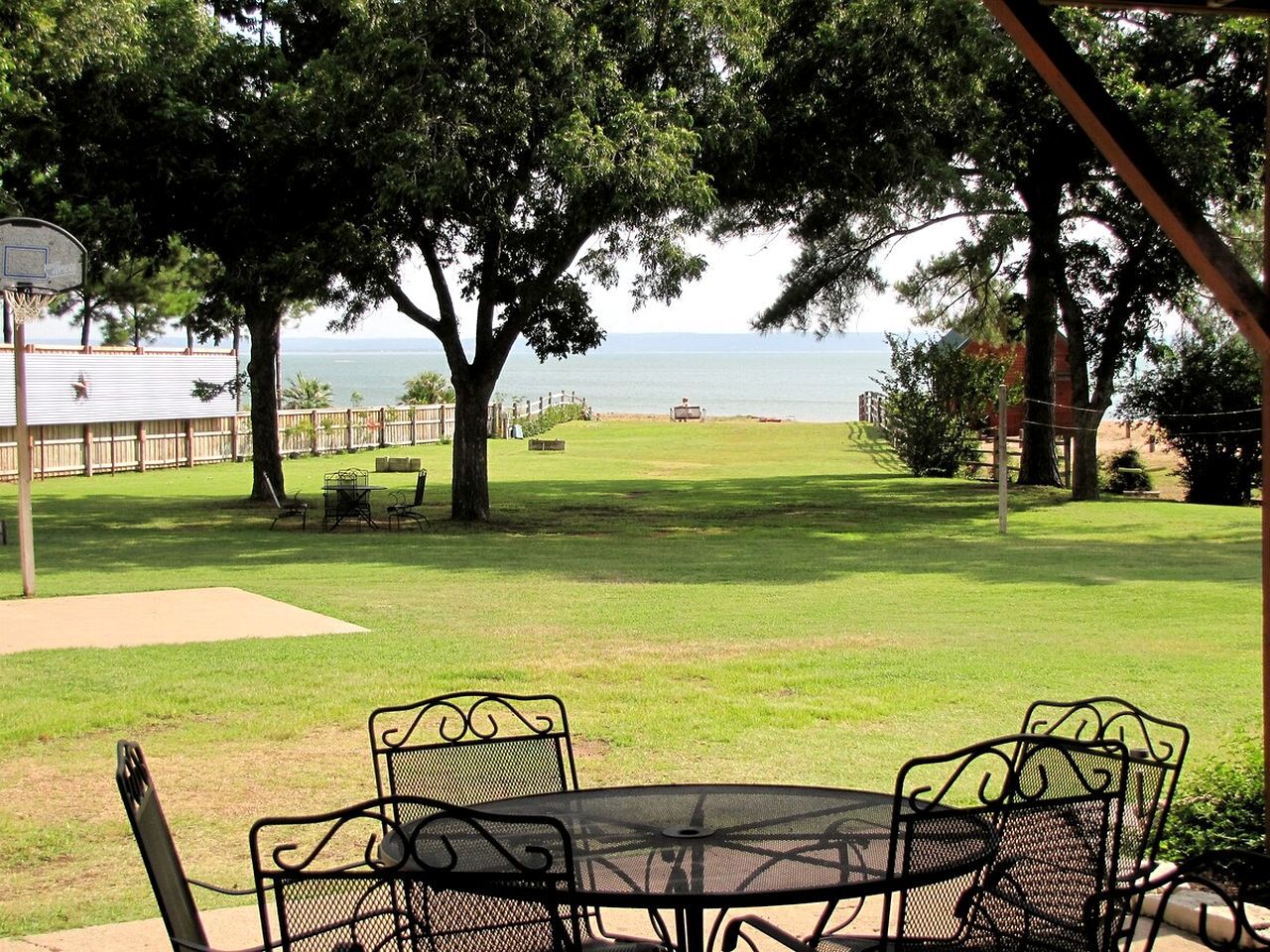 A patio with a table and chairs, overlooking a grassy area with a basketball hoop and a view of the sea.
