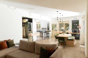 A modern living room with a sofa, a dining area with a table and chairs, a kitchen with cabinets and a window, and a hallway leading to another room. - Ria Oasis Residence, Luxury 3-Bedroom Apartment with Private Pool (Agios Nikolaos)