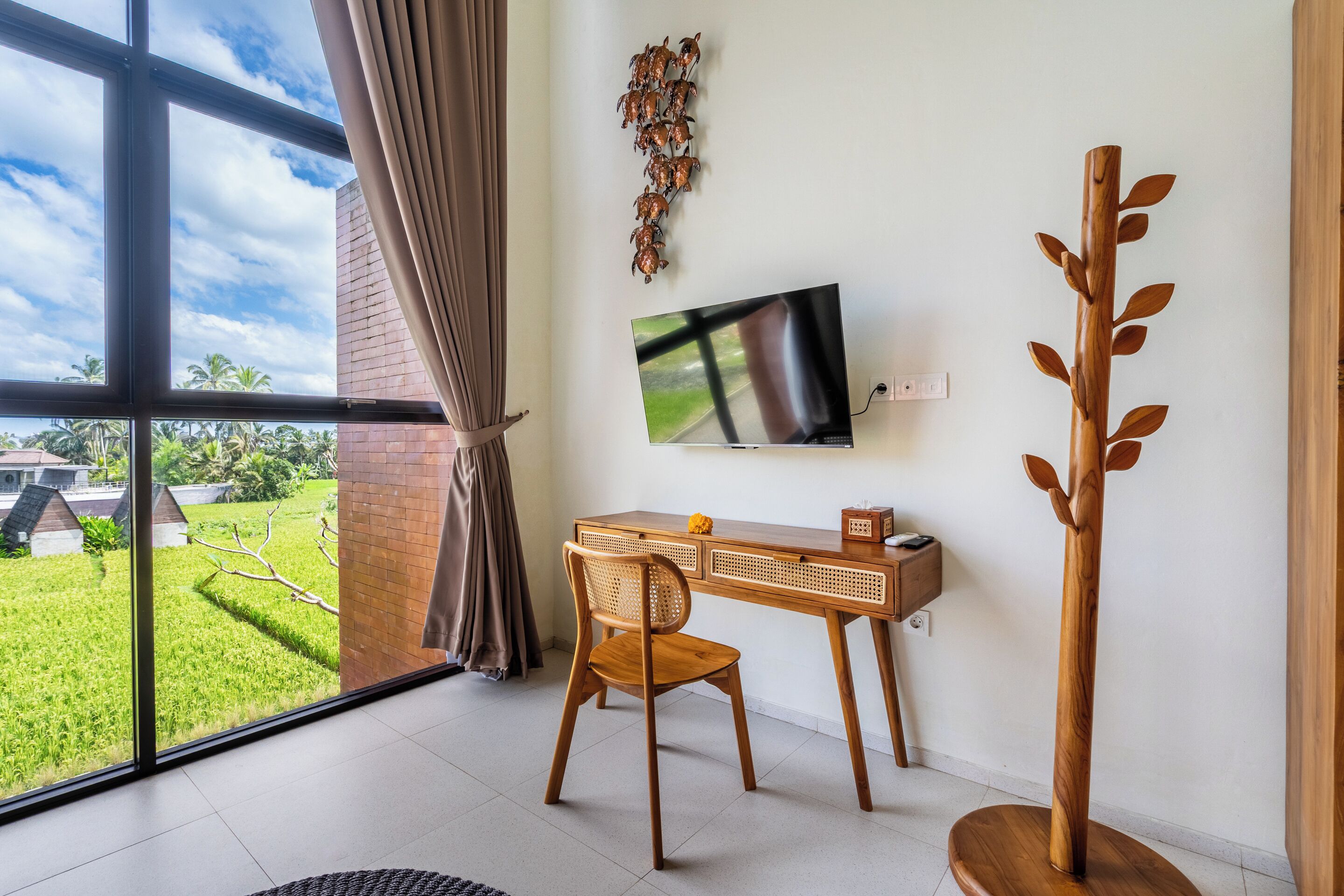 Een kamer met een bureau, stoel, televisie en uitzicht op een groen landschap door grote ramen.