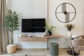 Ein modernes Wohnzimmer mit Flachbildfernseher, einer runden Wanduhr, einem Topf mit Pflanze, einem kleinen Tisch mit Vase und einem gepolsterten Hocker.