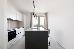A modern kitchen with a central island, built-in oven, and a view of the city through large windows. - 2 bedroom accommodation in OOSTENDE (OOSTENDE)