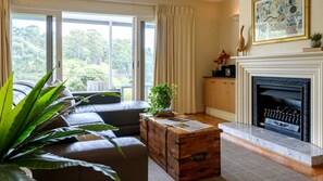 A living room with a sofa, a wooden chest, a fireplace, and a view of greenery outside. - Impressive Vacation Rental with Fireplace, Spa Bath and Wonderful Lake Views in Daylesford, Victoria (Daylesford)