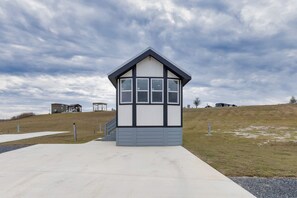 A small, single-story building with a sloped roof, a concrete pathway leading to it, and a grassy hill in the background. - Stylish and Comfortable Tiny Home Getaway with Resort-Style Amenities and Pool Near Fredericksburg, TX (Fredericksburg)