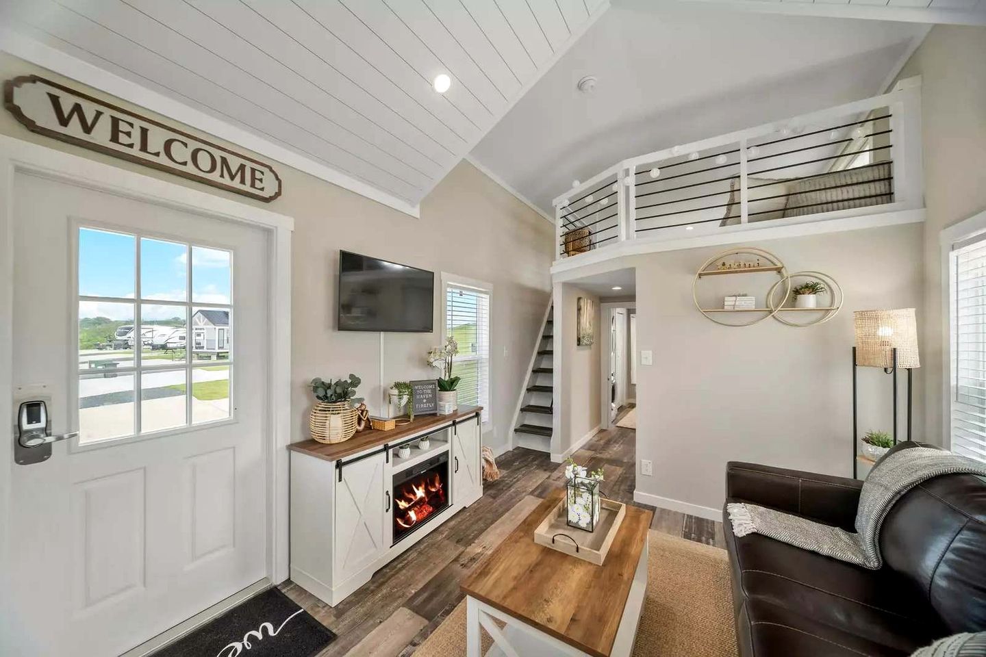 A spacious living area with a fireplace, a TV mounted on the wall, a staircase leading to an upper level, and a welcome sign on the door.