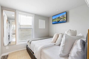 A bedroom with a bed, a large window with blinds, a flat-screen TV mounted on the wall, and a mirror. - Stylish and Comfortable Tiny Home Getaway with Resort-Style Amenities and Pool Near Fredericksburg, TX (Fredericksburg)